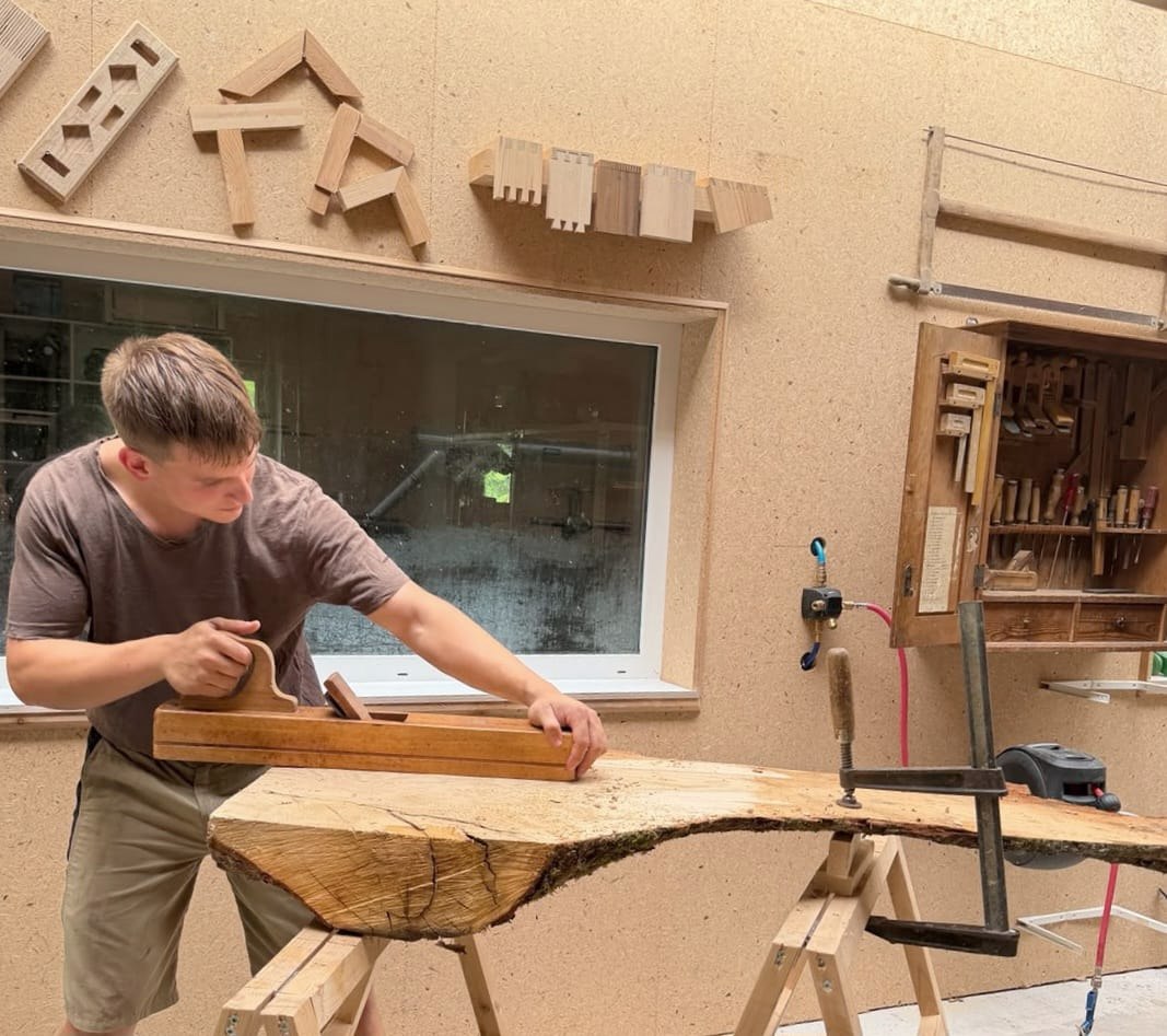 Noah Wolf hobelt in seiner Werkstatt eine Baumscheibe von Hand mit einem alten Holzhobel