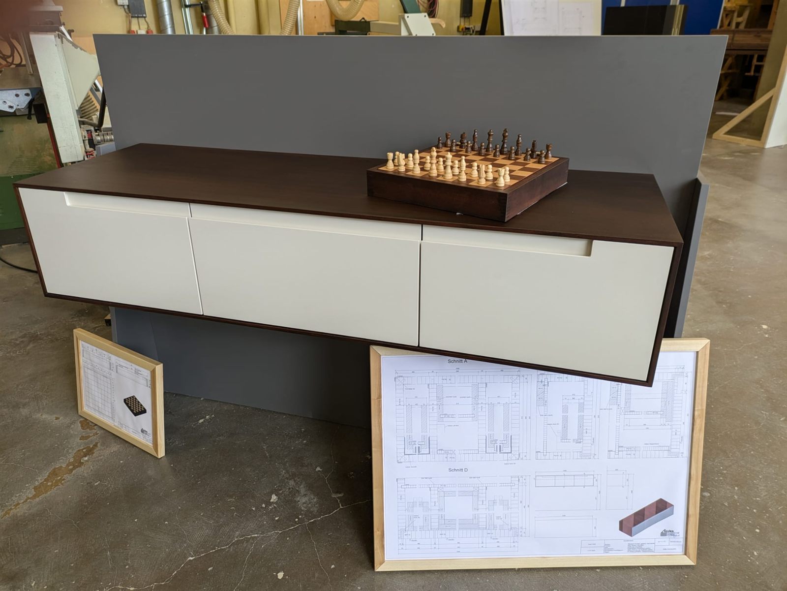 Hängendes Sideboard aus gebeiztem Mahagoni mit drei weißen Schubladen, darauf ein passendes Schachbrett, davor die gerahmten Konstruktionszeichnungen